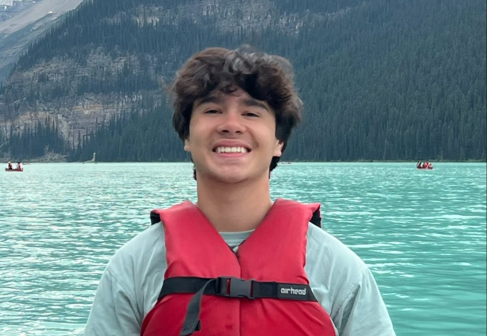 Young man smiling in a red life jacket on a turquoise lake with forested mountains in the background